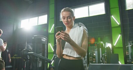 Strong athlete attractive woman is standing at the gym and resting after hard training. Girl is looking at smartphone screen while holding gadget in her hand. Stock video - Powered by Adobe