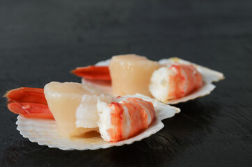 Raw scallop with a boiled shrimp tail in a shell on a black wooden surface.
