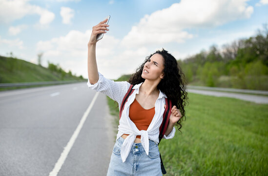 Upset young woman walking along road, raising hand with smartphone, looking for signal, having no connection, outdoors