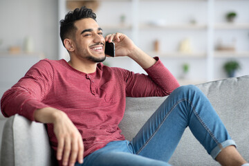 Happy middle-eastern guy sitting on couch, talking on phone - Powered by Adobe
