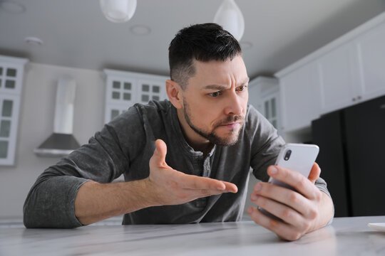 Emotional man with smartphone at table in kitchen. Online hate concept
