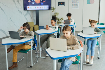 Diverse group of young children using laptops while sitting at desks in school classroom, copy space