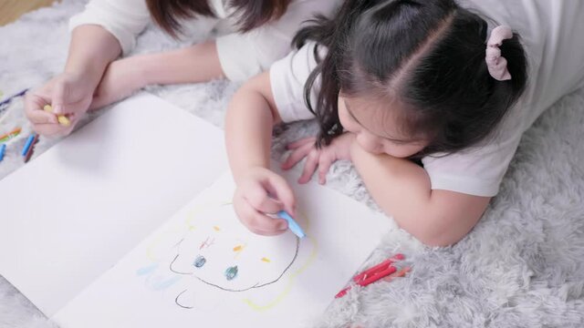 Top view of mom and kid lying on the floor doing painting by coloured pencils and talking together in living room with lots of happiness