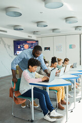 Vertical side view portrait of young boy using computer in IT class with male teacher helping him,...