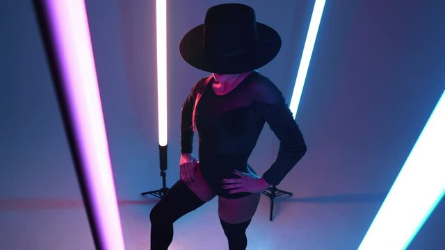 Sexy woman in hat posing flexibly between long glowing lamps, studio background. Femme fatale, stylish outfit of contemporary model dancer.