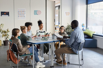 Fototapeta Full length shot of diverse group of children sitting at table with male teacher in modern school classroom obraz