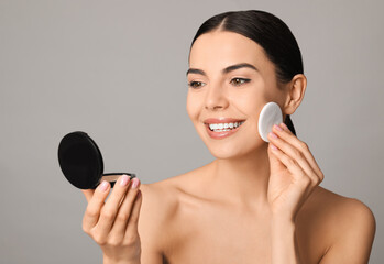 Beautiful young woman applying face powder with puff applicator on grey background - obrazy, fototapety, plakaty