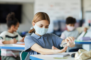 Portrait of young girl sanitizing hands in school classroom, covid safety measures, copy space