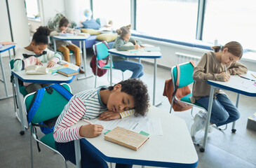 Obraz premium High angle portrait of mixed-raced boy sleeping at desk in school classroom, copy space