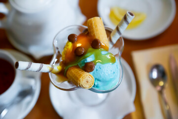 Fototapeta na wymiar Balls of yellow and blue ice cream with waffle tube in glass ramekin on table. Top view