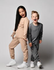 Kids in cosy cotton sportswear play together. Happy blond kid boy and brunette kid girl in pants and hoodies stands close to each other screaming and laughing over gray background