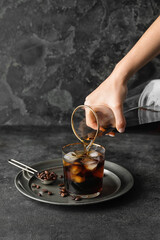 Pouring of cold brew coffee from jug into glass on grunge background
