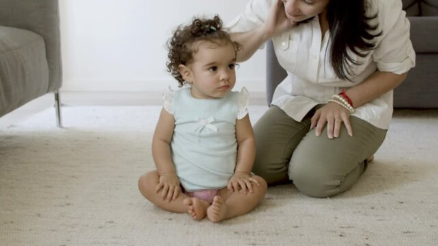 Serious Girl Sitting On Carpet With Mom At Home. Caucasian Mother Stroking Her Daughter On Head, Trying To Teach Her. Cute Baby Shaking Her Head With Displeasure. Motherhood, Development Concept.