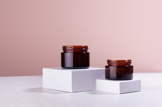Opened brown cosmetics containers for moisturizer cream on white podium with beige wall, mockup. Template for branding identity for cosmetics produce.