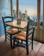 Empty restaurant cafe table and chairs near window with view of historic buildings and ocean at sunset