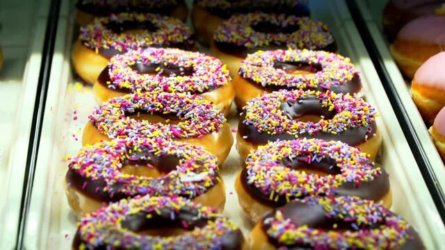 Krispy Kreme doughnuts under glass counter in shop. Sweat chocolate and sugar toppings. Cinematic slider shot.