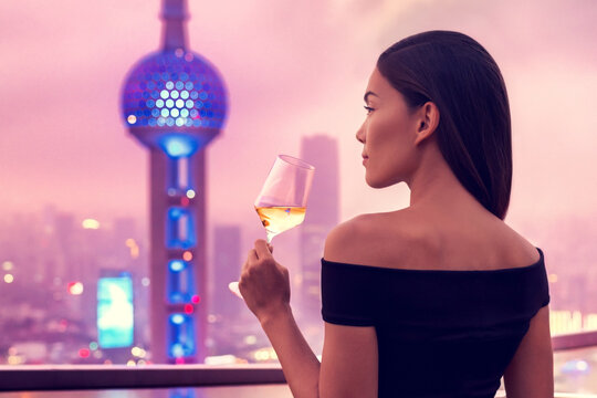 Elegant asian woman in gown drinking white wine glass at rooftop bar terrace looking at city lights skyline view of Shanghai in sunset. Luxury travel or high end lifestyle.