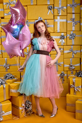 infantile playful female in colourful beautiful dress posing at camera, holding air balloons in hands, celebrating birthday, after party, receive many gifts boxes