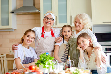 Friendly kind family having good time together at home in kitchen, smiling, after cooking, wearing aprons. Portrait of positive women and kids hugging each other, in domestic atmosphere