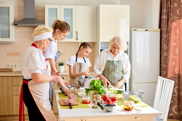 caucasian elderly grandparents cooking with granddaughters in the kitchen. two generation family spending quality time together. side view on family having talk during process of cooking