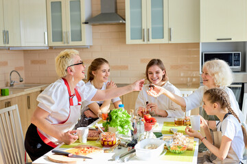 Joyous family women and children drinking tea after cokking, side view on cheerful females having rest, sitting at table enjoying time together, have talk, laughing. at home, in bright kitchen