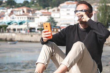 young man at the marina having coffee while using his mobile phone
