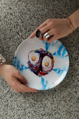 Ceramic decorative plate with colorful painting of modern French artist. Female hands with silver bracelets and rings are holding designer dish for home decor on the gray granite surface. 