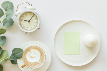 Blank paper card mockup and white alarm clock, coffee, candle, clean notebook, and flower on white background. Template for your design. Top view, flat lay. Minimal stationery scene.