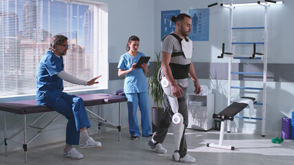 Man in exoskeleton learning to walk on treadmill near doctors - obrazy, fototapety, plakaty