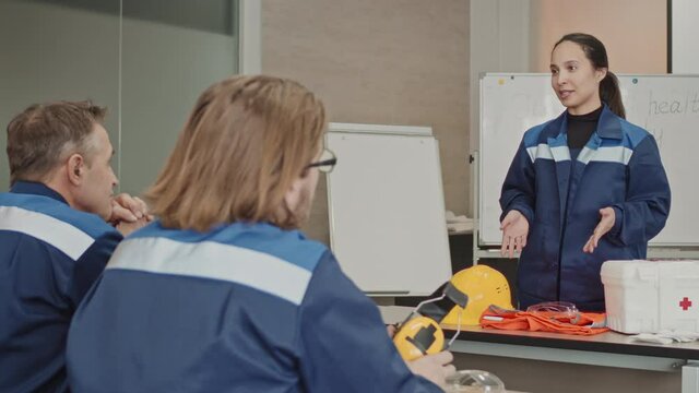 Medium PAN shot with slowmo of female plant supervisor giving lecture on first aid basics and safety rules at manufacturing facility to adult workmen sitting at desk in class