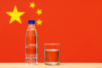 A bottle of clean drinking water and a glass stand on the table against the background of the flag of China. A concept for the supply of clean drinking water in China.