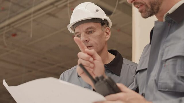 Low-angle chest-up of middle-aged Caucasian foreman wearing safety helmet, cropped constructor fighting with him over project documents