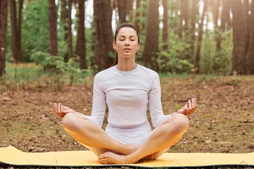 Fotobehang Front view of relaxed sporty girl with ponytail hairstyle sitting in lotus pose on mat, keeping eyes closed, legs crossed, yoga practicing in forest, woman wearing white sportswear. © sementsova321