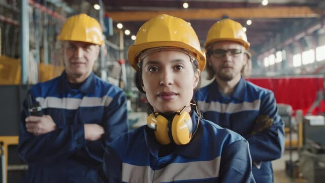 Portrait shot of Mixed-race female shift supervisor with noise-cancelling headphones and two men in hard hats and coveralls standing at factory and looking at camera