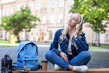 Portrait of a female college student sitting on a lawn using her smartphone at a sunny day. Caucasian high school student girl listening to music with her headphones on the phone in university park.