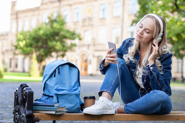 Positive happy cute student girl wearing denim jacket sitting on bench outdoors at campus, using mobile phone chatting. Blond young woman is listening music with headphones on university background.