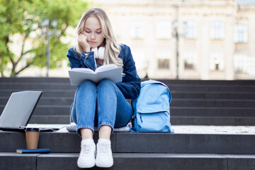 Tired young blond woman is sitting on stairs with laptop, student doing homework, preparing to pass exam, girl working on computer after sleepless night. High school lifestyle. Education concept.