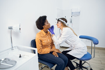 Fototapeta premium Pharyngitis and laryngitis treatment concept. Professional female doctor laryngologist, looking at throat of african american woman patient with wooden spatula in modern clinic