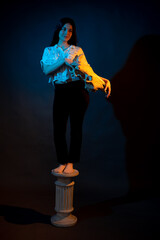 A girl stands on a Greek column with a bull skull in her hands illuminated by blue and yellow light