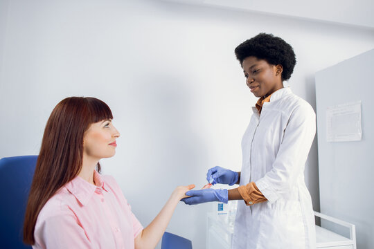 Female African American Lab Technician, Drawing Finger Blood For Test. Young Caucasian Lady Patient Trusting Her Doctor And Having Blood Test