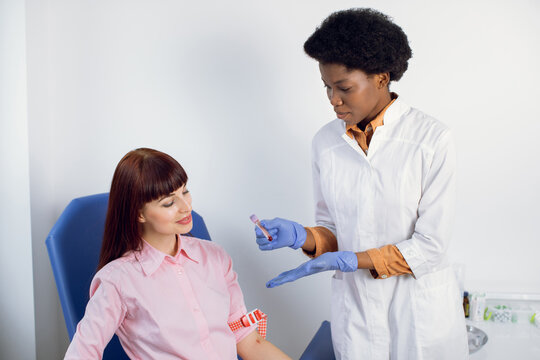 Blood Test In Modern Medical Laboratory. Young Smiling Caucasian Woman Patient, Sitting At Modern Lab In The Chair, While Confident Female Black Doctor Showing Test Tube With Blood Sample