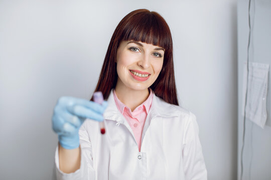 Female Caucasian doctor scientist examining a chemical reaction or blood in a test tube, posing with smile to camera in modern clinical laboraory. Focus on face of doctor