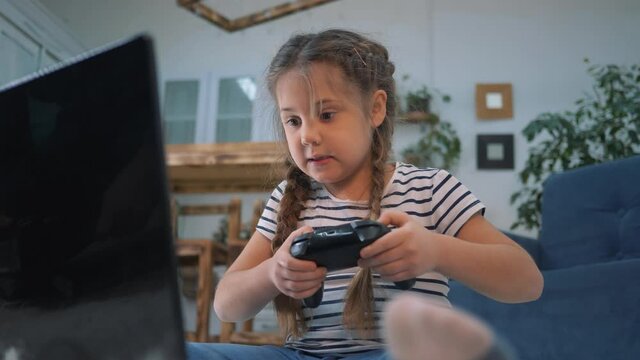 Happy girl playing video games with a joystick. Child gambling addiction. Happy little girl playing video game home. Concept of child gambling addiction. Happy little girl learning to work on laptop
