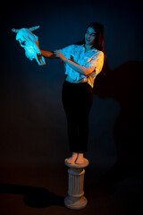 A girl stands on a Greek column with a bull skull in her hands illuminated by blue and yellow light
