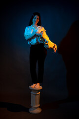 A girl stands on a Greek column with a bull skull in her hands illuminated by blue and yellow light