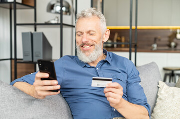 Happy optimistic mature man holding smartphone and credit card, paying bills online, senior male...