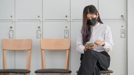 An attractive young woman wearing a face mask and using her cellphone while sitting on a chair in a waiting room