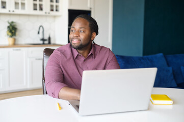 Serene African-American businessman using laptop for remote work, sitting at the desk and looks away, waiting for inspiration and news ideas, positive black guy typing sitting in the kitchen