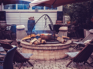 A cauldron of vegetables hangs over a stone well with wood and sun loungers around outdoors. Street cafe
