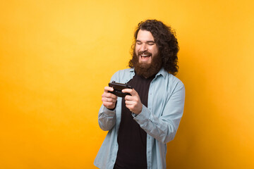 Photo of cheerful young bearded man playing games at smartphone over yellow background